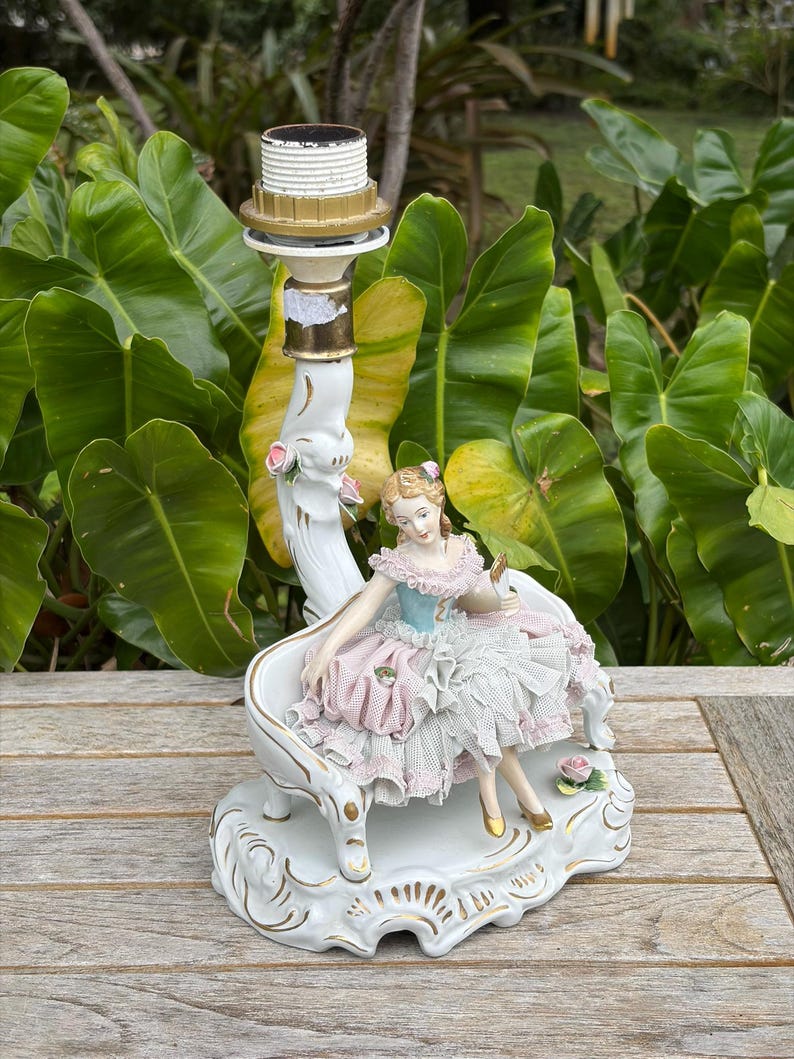May include: A white porcelain lamp base featuring a seated figurine in a ruffled dress. The figurine is in shades of pink, blue, and white, with gold accents. The lamp base has a decorative design and a brass lamp holder.