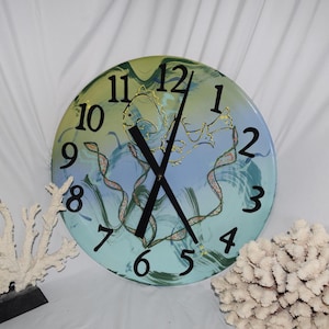 May include: A round wall clock with a sea-themed design. The clock face features a blue and green background with black numerals and hands. Decorative coral accents are placed on either side of the clock.