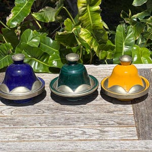 May include: Three colorful ceramic tagine pots with silver-colored metal trim. The tagines are blue, green, and yellow. The pots are sitting on a weathered wooden surface, with green foliage in the background.