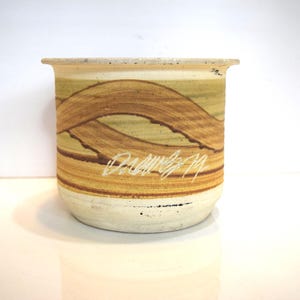 May include: A ceramic pot with a cream-colored base and a tan rim. The pot features a design of brown, wavy lines over a yellow-green background. The text "Dallos 79" is inscribed in white.