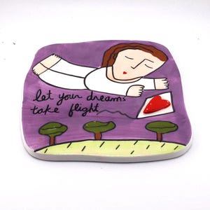 May include: A hand-painted ceramic piece with a purple background features a person in white clothing dreaming. The text "let your dreams take flight" is written in cursive. A red heart and three green trees are also depicted.