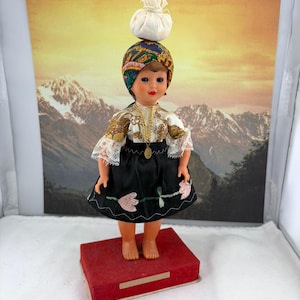Könnte beinhalten: Eine Vintage-Puppe in traditioneller Kleidung, die auf einem roten Sockel steht. Die Puppe trägt einen schwarzen Rock mit rosa Blumenstickerei, eine weiße Bluse mit Spitzärmeln und ein gemustertes Kopftuch. Ein kleines weißes Bündel balanciert auf dem Kopf der Puppe.