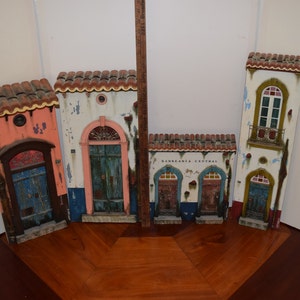 May include: Five miniature wooden buildings with colorful facades and doors. The buildings are painted in shades of pink, white, blue, green, and brown. The building in the center has the text "BARBEARIA CENTRAL" on the front.