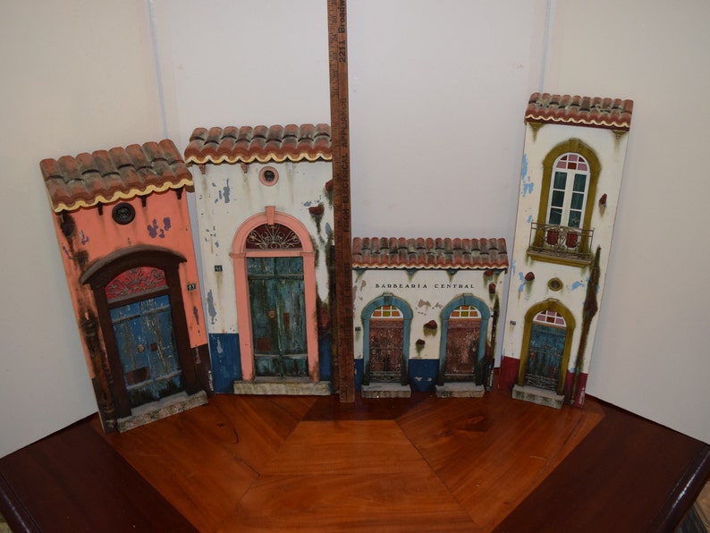 May include: Five miniature wooden buildings with colorful facades and doors. The buildings are painted in shades of pink, white, blue, green, and brown. The building in the center has the text "BARBEARIA CENTRAL" on the front.