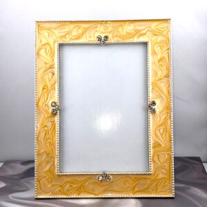 May include: A rectangular photo frame with a yellow and cream swirl pattern. The frame has a silver border and is adorned with three sets of three small, clear crystals. The frame is designed to hold a photo.