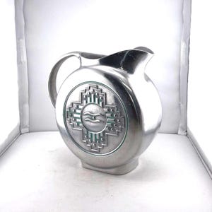 May include: A silver-colored pitcher with a rounded body and a handle. The pitcher features a decorative emblem with a cross-like design and a central water wave symbol. The pitcher is made of metal and has a spout for pouring.