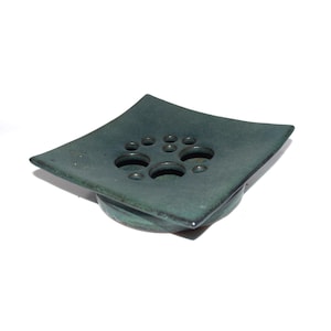 May include: A square, teal-colored ceramic flower frog with multiple circular holes for arranging stems. The frog has a slightly raised base and a glossy finish. The design is modern and minimalist, suitable for floral arrangements.