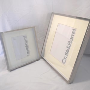 May include: Two silver-toned picture frames from Crate & Barrel. One frame is square, the other is rectangular. Both have a white mat and a clear glass cover. The frames are displayed on a white surface.