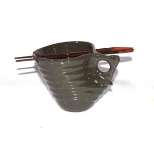 May include: Olive green ceramic ramen bowl with a unique handle and ridged design. Includes a pair of wooden chopsticks resting across the top. The bowl is designed for serving Asian cuisine.