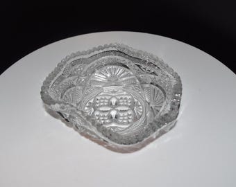May include: A clear pressed glass bowl featuring a square shape with rounded corners and a scalloped, beaded rim. The exterior surface displays intricate geometric patterns, including fan-like motifs and a central cross-hatched diamond design.