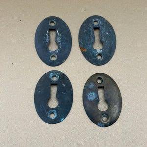 May include: Four oval-shaped metal keyhole escutcheons with a dark, weathered patina. Each plate features a central keyhole cutout and two small circular screw holes at the top and bottom, showing signs of oxidation and surface wear throughout.