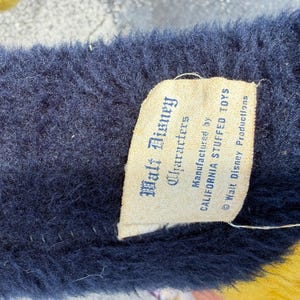 May include: Close-up of a plush toy's tag. The tag reads "Walt Disney Characters" and "CALIFORNIA STUFFED TOYS." The plush has a dark blue, fuzzy texture. The tag is cream-colored with black text. The bottom of the plush is yellow.