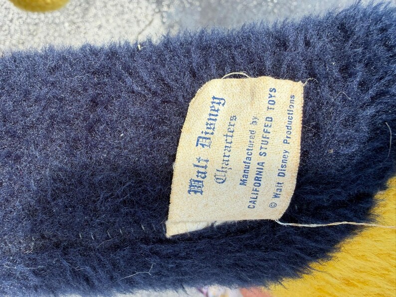 May include: Close-up of a plush toy's tag. The tag reads "Walt Disney Characters" and "CALIFORNIA STUFFED TOYS." The plush has a dark blue, fuzzy texture. The tag is cream-colored with black text. The bottom of the plush is yellow.