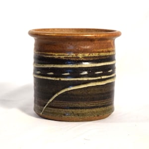 May include: A handmade ceramic jar with a cylindrical shape. The jar features a brown rim and base, with a dark brown and black body. White and cream-colored horizontal lines and a curved accent adorn the body, creating a unique, rustic aesthetic.