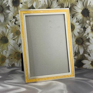 May include: A rectangular picture frame featuring a smooth, glossy yellow border with a marbled effect, accented by a thin, silver-toned inner trim with a beaded texture. The frame includes a neutral gray insert and a black easel-style back support.