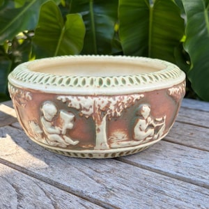 May include: A round, antique ceramic planter with a detailed relief design. The planter features a cream-colored rim with a pierced pattern, and a brown body with figures and a tree. The planter is sitting on a wooden surface.