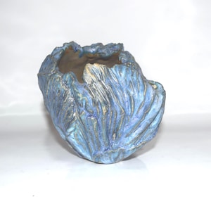 May include: A handcrafted ceramic vase with a textured, organic design. The vase features a blend of blue and gold hues, with a rough, uneven surface. The open top and unique shape make it a decorative piece.