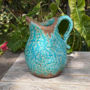 May include: A turquoise ceramic pitcher with a textured, crackled glaze. The rim and base are a contrasting brown. The pitcher has a curved handle and a rounded body. The background features green foliage.