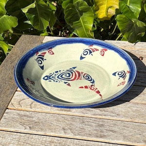 May include: Ceramic serving bowl with a blue rim and a cream-colored interior. The bowl is decorated with stylized red and blue fish designs. The bowl is sitting on a wooden table.
