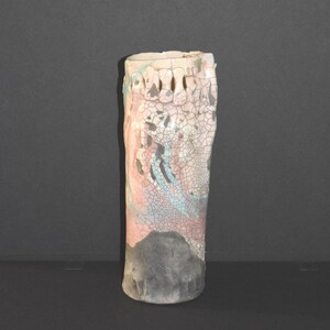 May include: A cylindrical ceramic vase featuring a crackle glaze finish in shades of pale pink, light blue, and grey. The upper rim displays a series of small, vertical indentations, while the base is finished in a solid, dark matte grey tone.