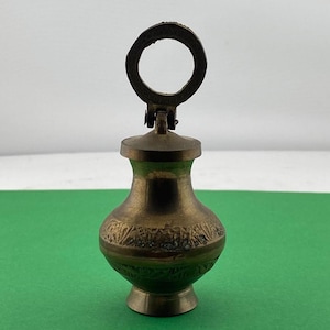 May include: A small, antique brass vase with a decorative floral pattern around the body. The vase has a lid with a circular handle. The vase is on a green surface.