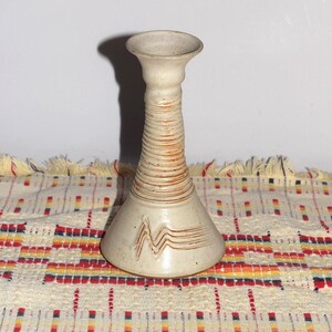 May include: A beige ceramic candlestick holder with a flared top and base. The neck features horizontal ribbed detailing, while the base displays a carved zigzag pattern. The ceramic has a matte finish with subtle brown accents within the grooves.
