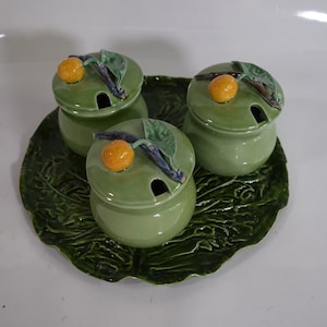 May include: Three green ceramic condiment jars with lids decorated with a small orange and a leaf, sitting on a green ceramic tray shaped like a cabbage leaf.