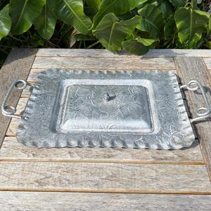 May include: A rectangular, silver-colored metal serving tray with a floral pattern and two handles. The tray has a scalloped edge and a central, lidded compartment. The tray is sitting on a wooden table.