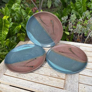 May include: Three ceramic plates with a two-tone design. Each plate is divided diagonally, with a blue-green section and a reddish-brown section. Black abstract lines decorate the reddish-brown side. The plates are on a wooden surface.