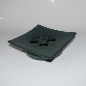 May include: A dark teal ceramic flower frog with a square top and multiple holes for arranging stems. The frog sits on a circular base, perfect for floral arrangements. The glaze has a matte finish.