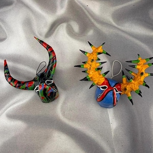 Könnte beinhalten: Zwei dekorative Hängeornamente. Das linke Ornament ist eine schwarz-rot-grüne Maske in Stierform mit geschwungenen Hörnern. Das rechte Ornament ist eine blau-rot-gelbe Maske mit zwei großen, stacheligen, gelben geweihartigen Strukturen.