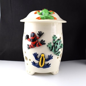 May include: A cream-colored ceramic jar with a lid, adorned with colorful frog figurines. The frogs are in various poses, with red, black, green, blue, and yellow accents. The jar has small circular cutouts and is elevated on three small feet.