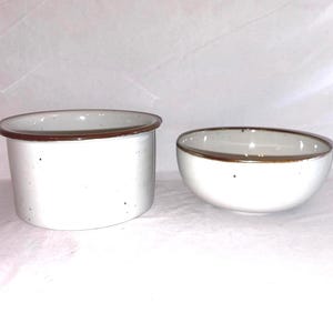 May include: A set of two ceramic kitchen vessels featuring a speckled off-white glaze and a solid brown rim. The first piece is a deep, straight-sided cylindrical container, while the second piece is a shallower, rounded bowl with curved sides.
