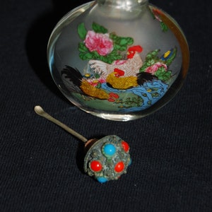 May include: A clear glass bottle with a silver stopper and a painted design of two roosters and flowers. The bottle is round and has a textured surface.