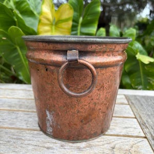 May include: A round copper bucket with a weathered, mottled patina and a dark metallic rim. The bucket features a single circular copper ring handle attached to the side with a flat metal bracket, showcasing a textured, aged surface finish.