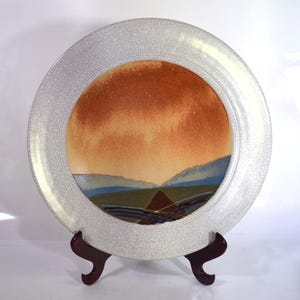 May include: A round ceramic plate featuring a white crackle glaze border and a central landscape illustration. The artwork depicts a warm orange sky above blue hills and a dark brown foreground, creating a scenic view on the smooth surface.