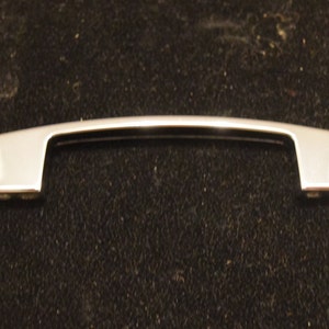 May include: A silver-toned cabinet pull with a curved design. The pull has a rectangular shape with rounded edges.