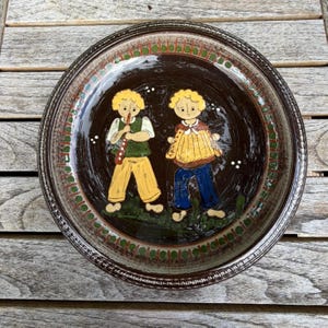 May include: A round, decorative plate with a dark brown center featuring two stylized figures. One figure plays a flute, the other an accordion. The rim has a green and brown pattern. The plate is likely ceramic.