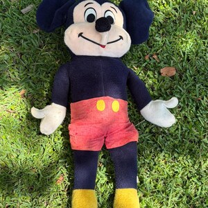 May include: A vintage Mickey Mouse plush toy, featuring a black body, large ears, and a cream-colored face. Mickey wears red shorts with yellow buttons, white gloves, and yellow shoes. The plush is lying on green grass.