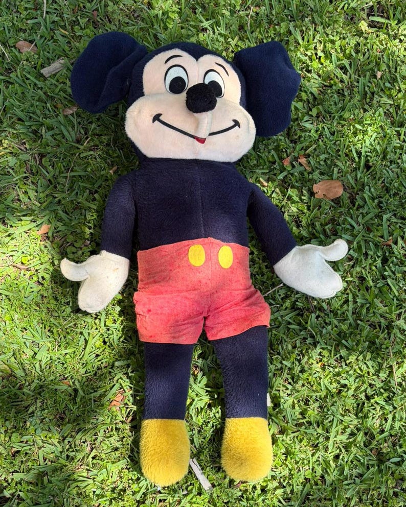 May include: A vintage Mickey Mouse plush toy, featuring a black body, large ears, and a cream-colored face. Mickey wears red shorts with yellow buttons, white gloves, and yellow shoes. The plush is lying on green grass.