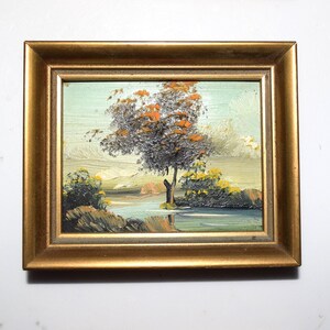 Op de afbeelding: Een ingelijst olieverfschilderij toont een landschap met een boom met oranje en grijze bladeren. Het kunstwerk, tegen een lichtblauwe lucht, omvat water en groene struiken. Het schilderij zit in een goudkleurige houten lijst.