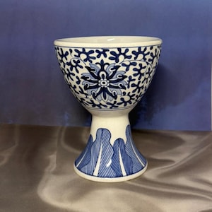 May include: A white ceramic egg cup with blue floral patterns. The cup has a wide top, a narrow stem, and a flared base. The blue design includes a central flower and leaf-like patterns. The egg cup is a decorative item.
