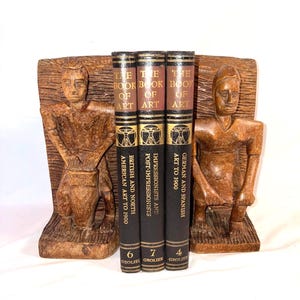 May include: A set of three vintage art books titled "The Book of Art" with brown and black covers, standing between two carved wooden bookends. The bookends feature a carved figure. The books are numbered 4, 6, and 7.