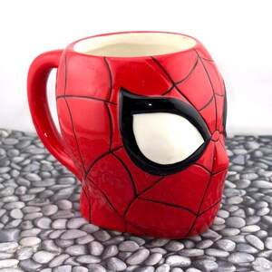 May include: A red ceramic mug shaped like Spider-Man's mask, featuring black web lines and a white eye. The mug has a red handle and a white interior. The mug is a collectible item.