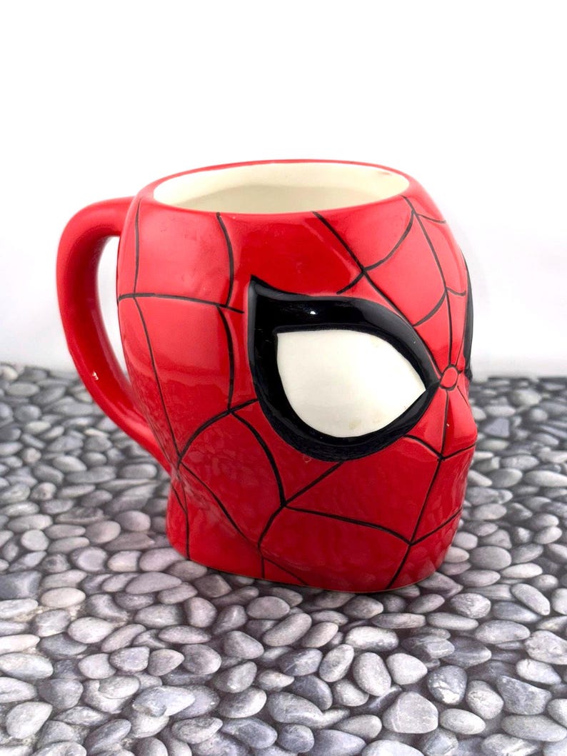 May include: A red ceramic mug shaped like Spider-Man's mask, featuring black web lines and a white eye. The mug has a red handle and a white interior. The mug is a collectible item.