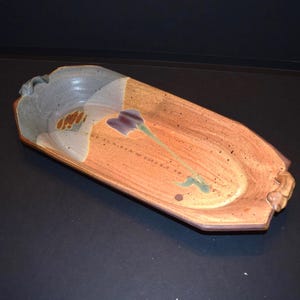 May include: An elongated, octagonal ceramic serving tray with a speckled terracotta-colored base. The interior features a painted purple flower with a green stem and a contrasting light gray glazed section at one end, finished with small handles.