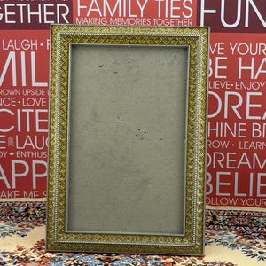 May include: A rectangular picture frame with a metallic gold finish and a textured, repeating geometric diamond pattern along the border. The frame features a thin, beaded inner edge and a plain, light gray backing board centered within the frame.