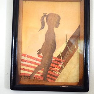 May include: A framed wall art piece featuring a dark brown silhouette of a person with a ponytail. The figure stands against a background of horizontal red and cream stripes and a textured tan area. The frame is black with a metallic hanging hook.