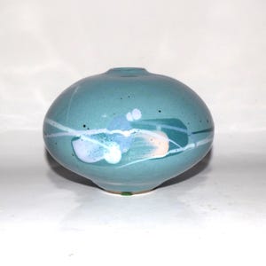 May include: A round, teal ceramic vase with a smooth, glossy finish. The surface features abstract, painted accents in shades of light blue, white, and soft peach, along with thin, horizontal white lines and small, dark speckles across the body.