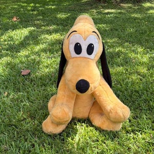 May include: A plush toy of Pluto, a yellow dog with black ears and a black nose, sits on green grass. The toy has large white eyes with black pupils and is a popular character from Disney.